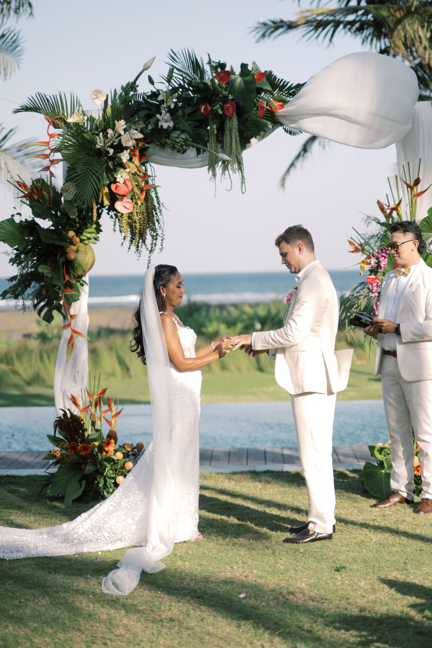 Bride and groom exchange vows under a tropical floral arch with ocean views at sunset.