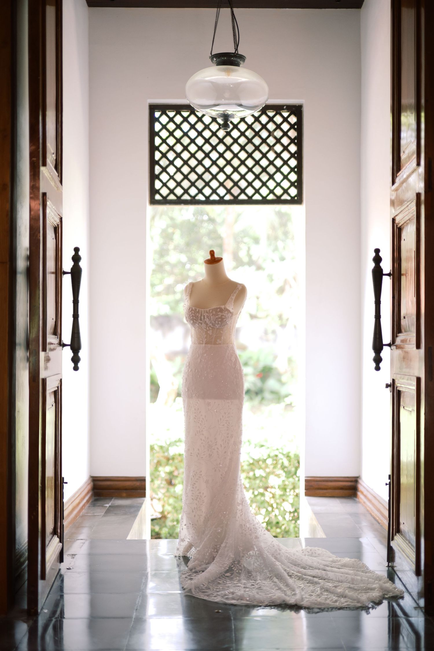 Elegant white lace wedding gown on a mannequin displayed in a bright doorway with lattice window.