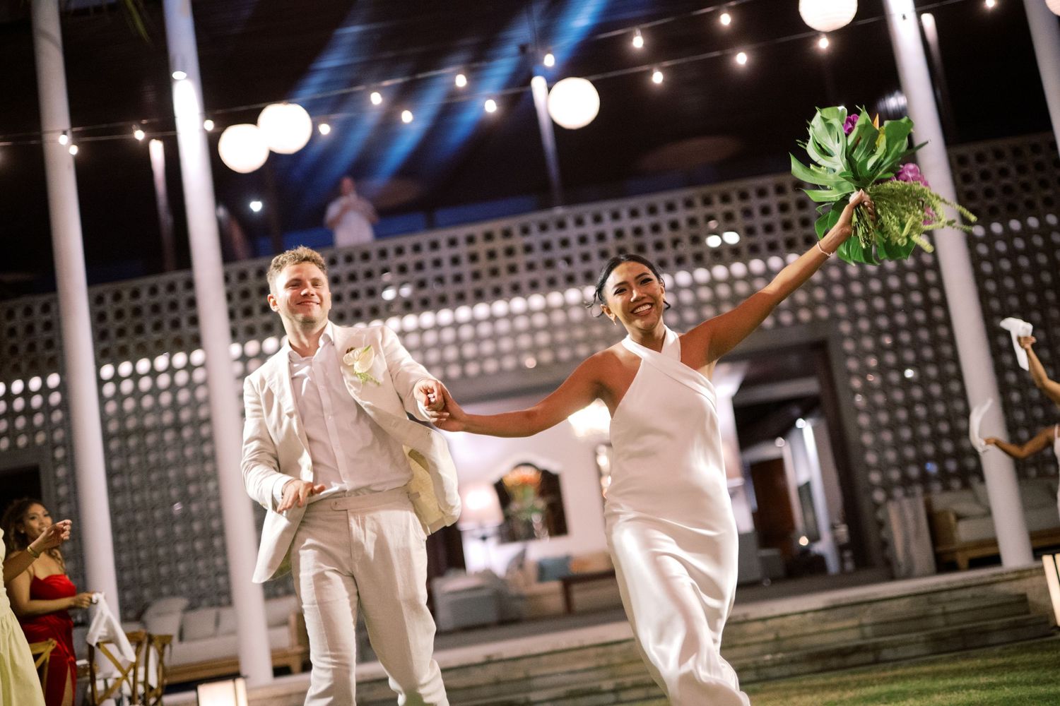 Bride in white jumpsuit raises bouquet triumphantly as groom in cream suit enters outdoor tropical wedding reception.