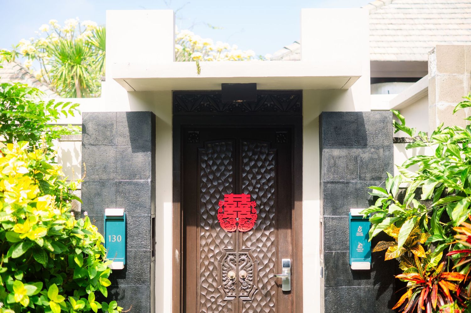 Ornate carved wooden entrance door with red Chinese double happiness symbol, framed by dark stone walls and lush tropical plants.