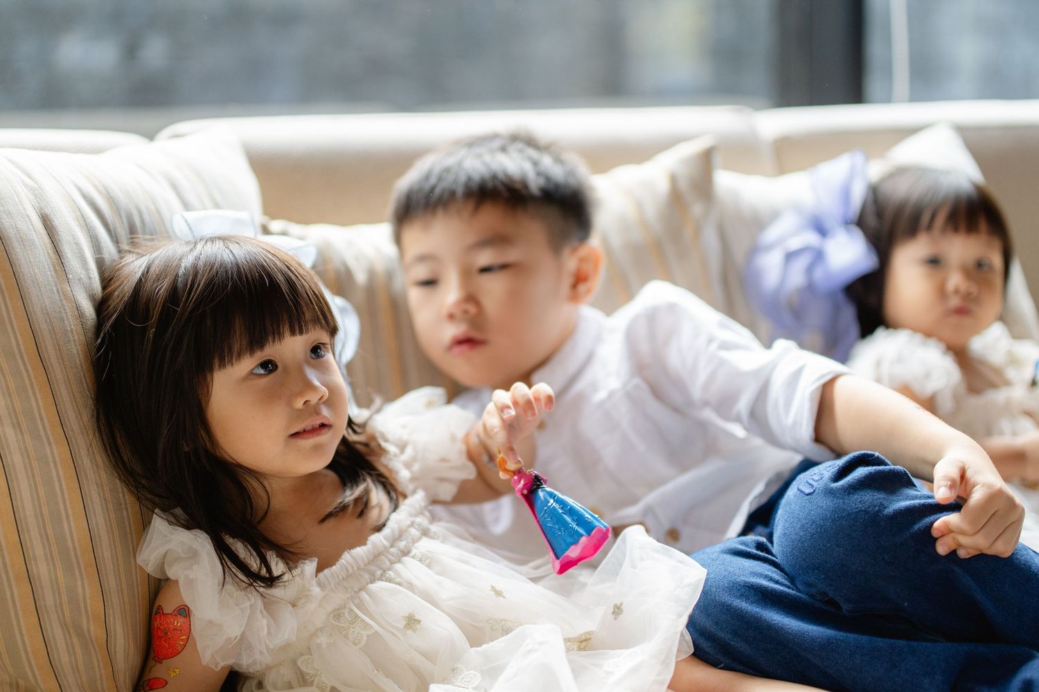 Three young Asian children relaxing together on a beige sofa, dressed in white and blue outfits.