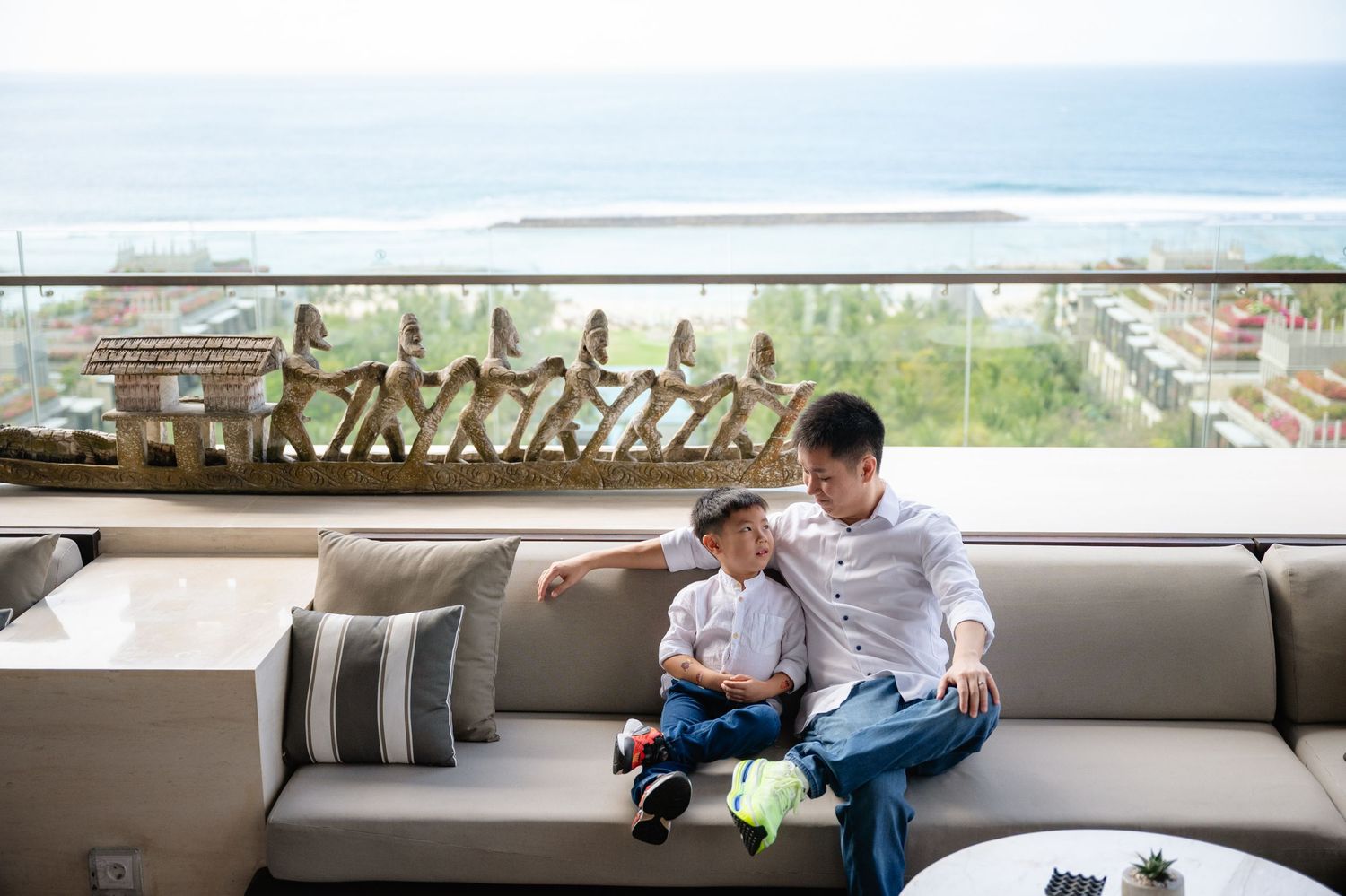 A father and young child sit together on an outdoor sofa with a scenic ocean view in the background.
