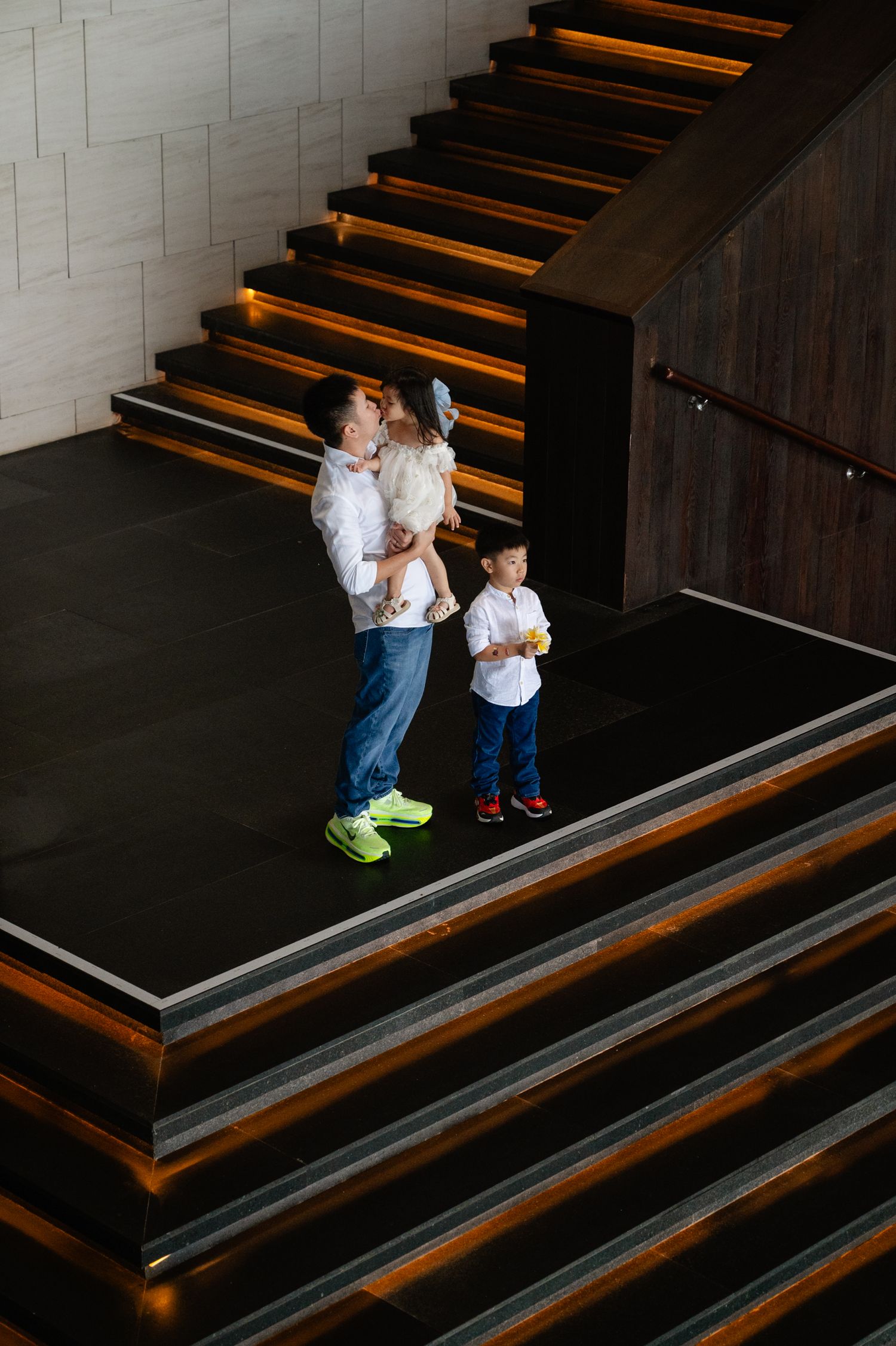 A man holding a toddler stands with a young boy on illuminated modern stairs in a contemporary building.