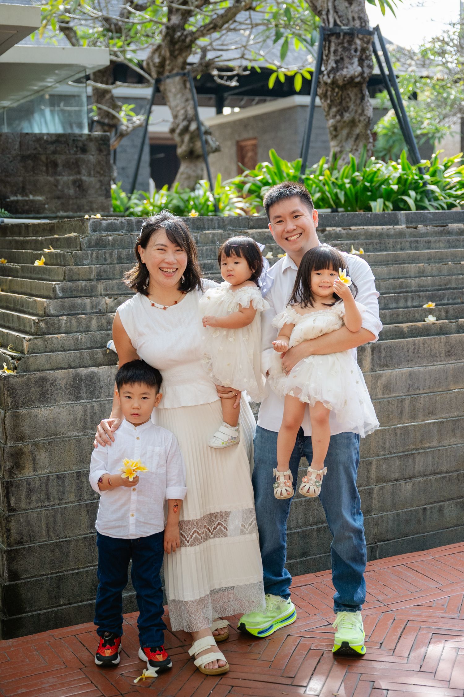 Happy Asian family of five posing together outdoors, dressed in white outfits, with two children being held by parents.