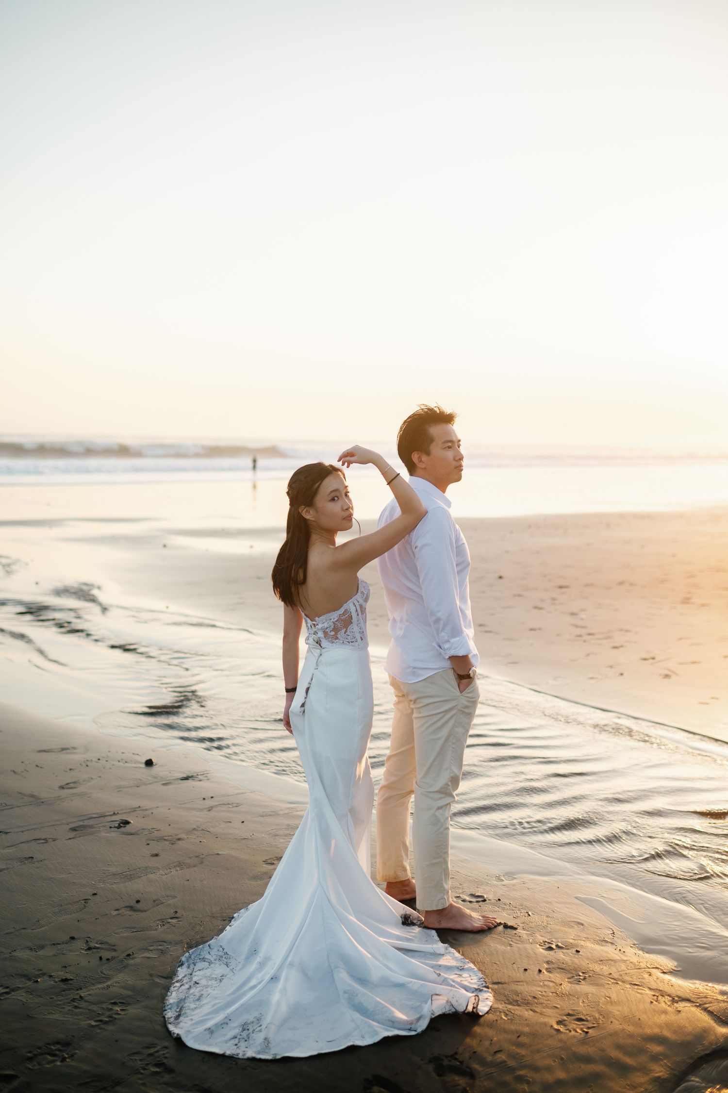 Couple in white wedding attire posing on a beach at sunset, woman in elegant gown touching man's hair.