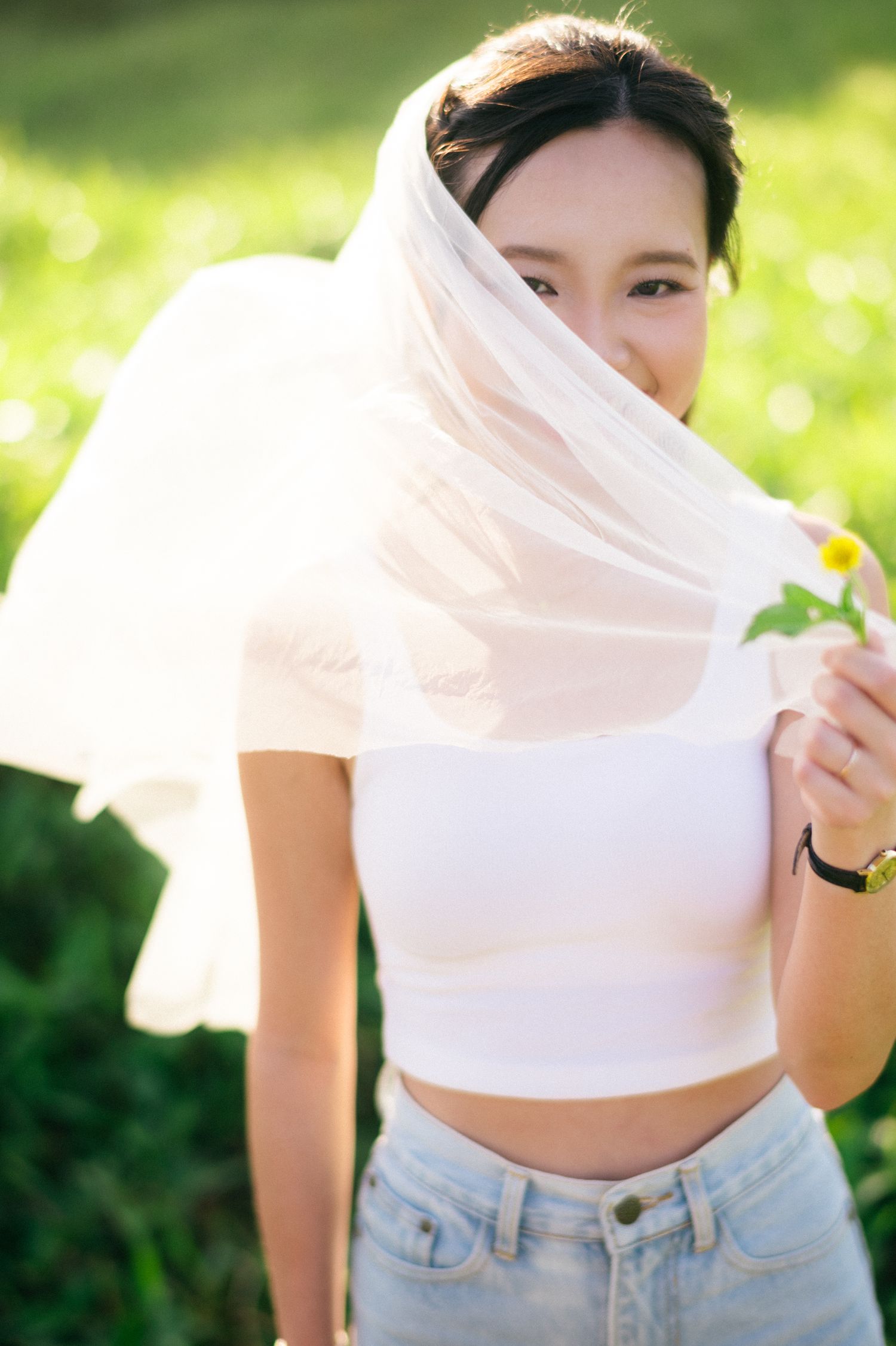 A young Asian bride in white crop top and veil holds a daisy flower while posing in a sunny green field.