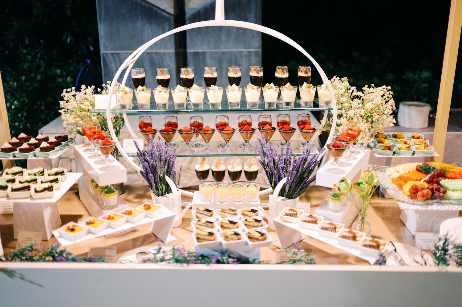 Elegant wedding dessert table display with tiered stands, chocolates, fruit tarts, and floral arrangements.