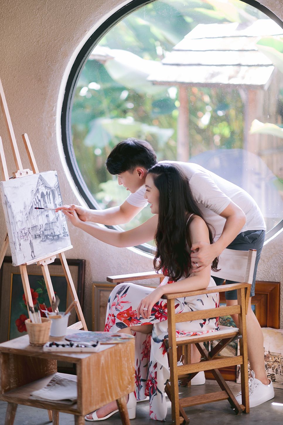 A couple shares a romantic moment painting together on an easel near a circular window in a cozy art studio.