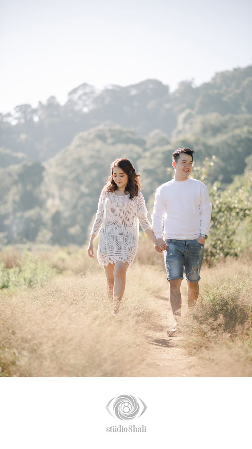 Couple holding hands walking on a sunny dirt path surrounded by golden grass and trees in a romantic outdoor photoshoot.