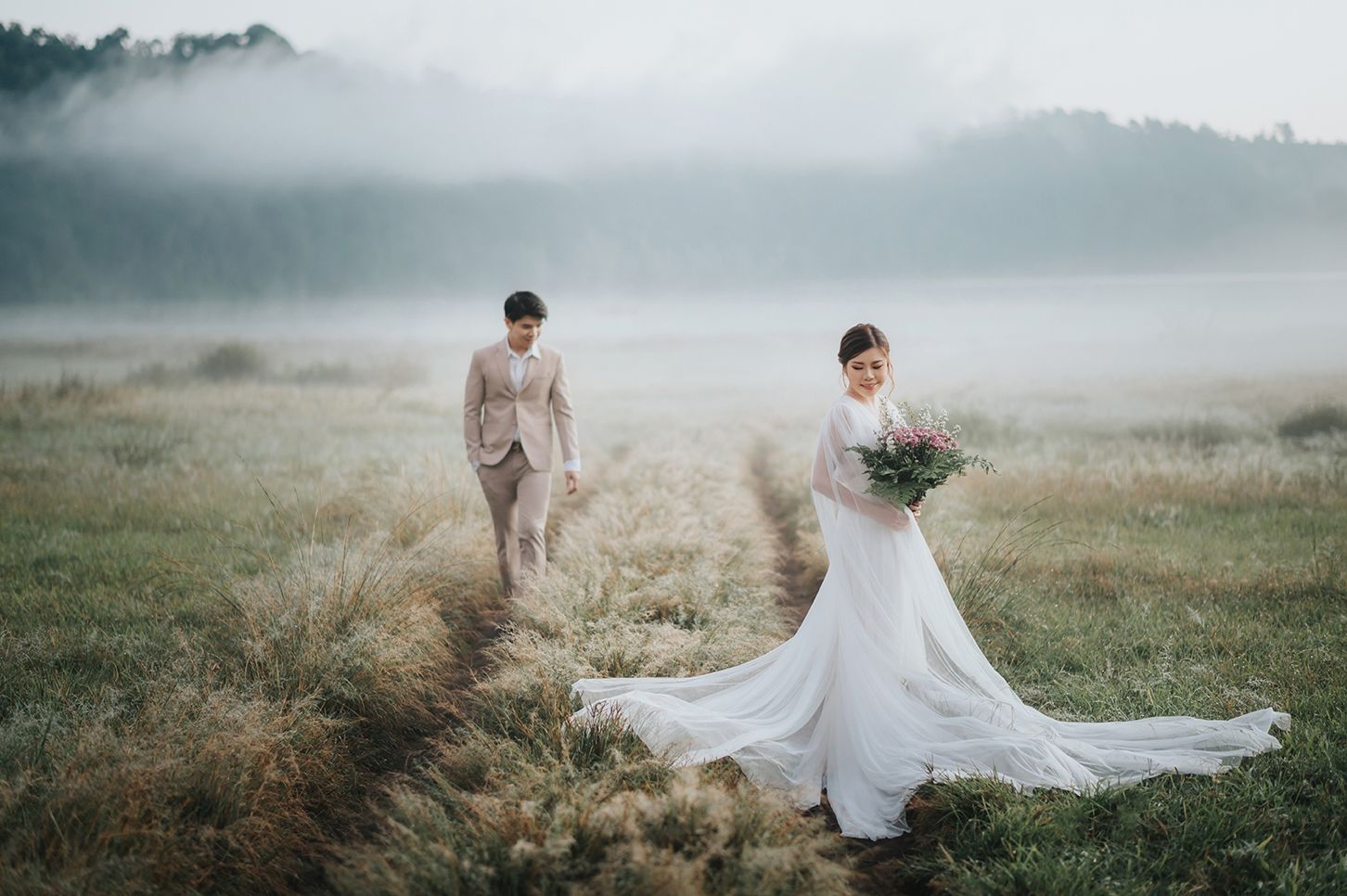 Bride in a flowing gown and groom in a tan suit walking through a misty meadow at dawn.