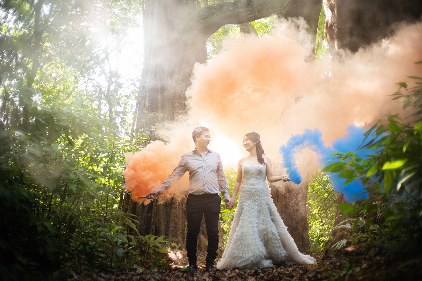 Couple poses in lush forest with colorful orange and blue smoke bombs for a dreamy wedding photoshoot.