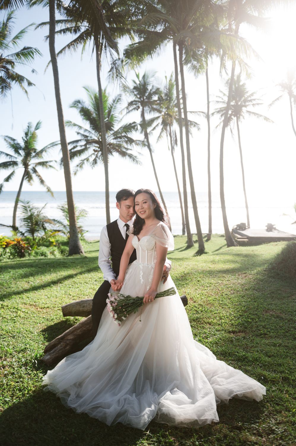 Couple in wedding attire poses among palm trees near beach, bride in flowing gown holding bouquet, groom in vest and tie.
