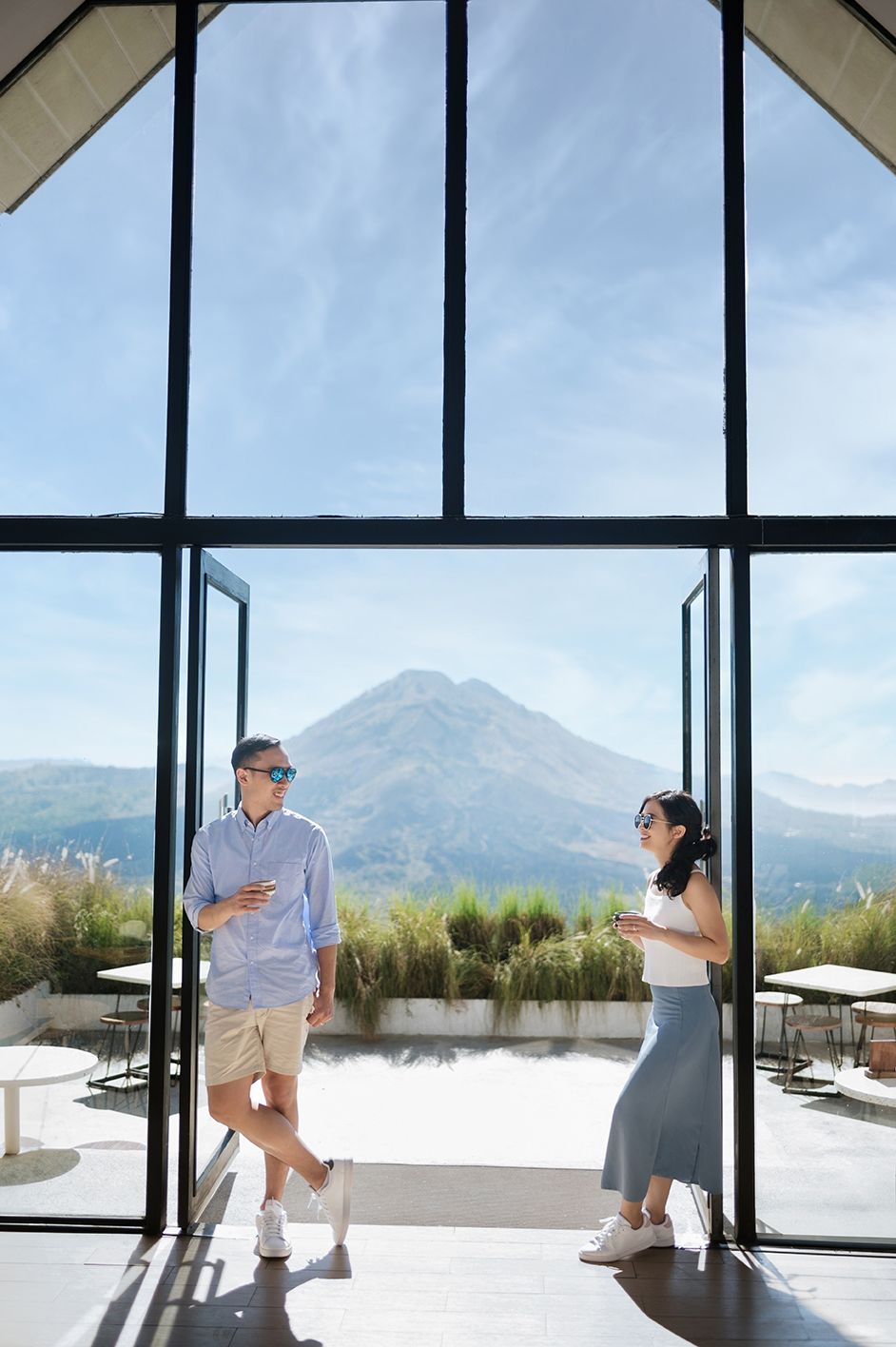 Couple standing in modern glass doorway with stunning volcanic mountain landscape backdrop on a sunny day.