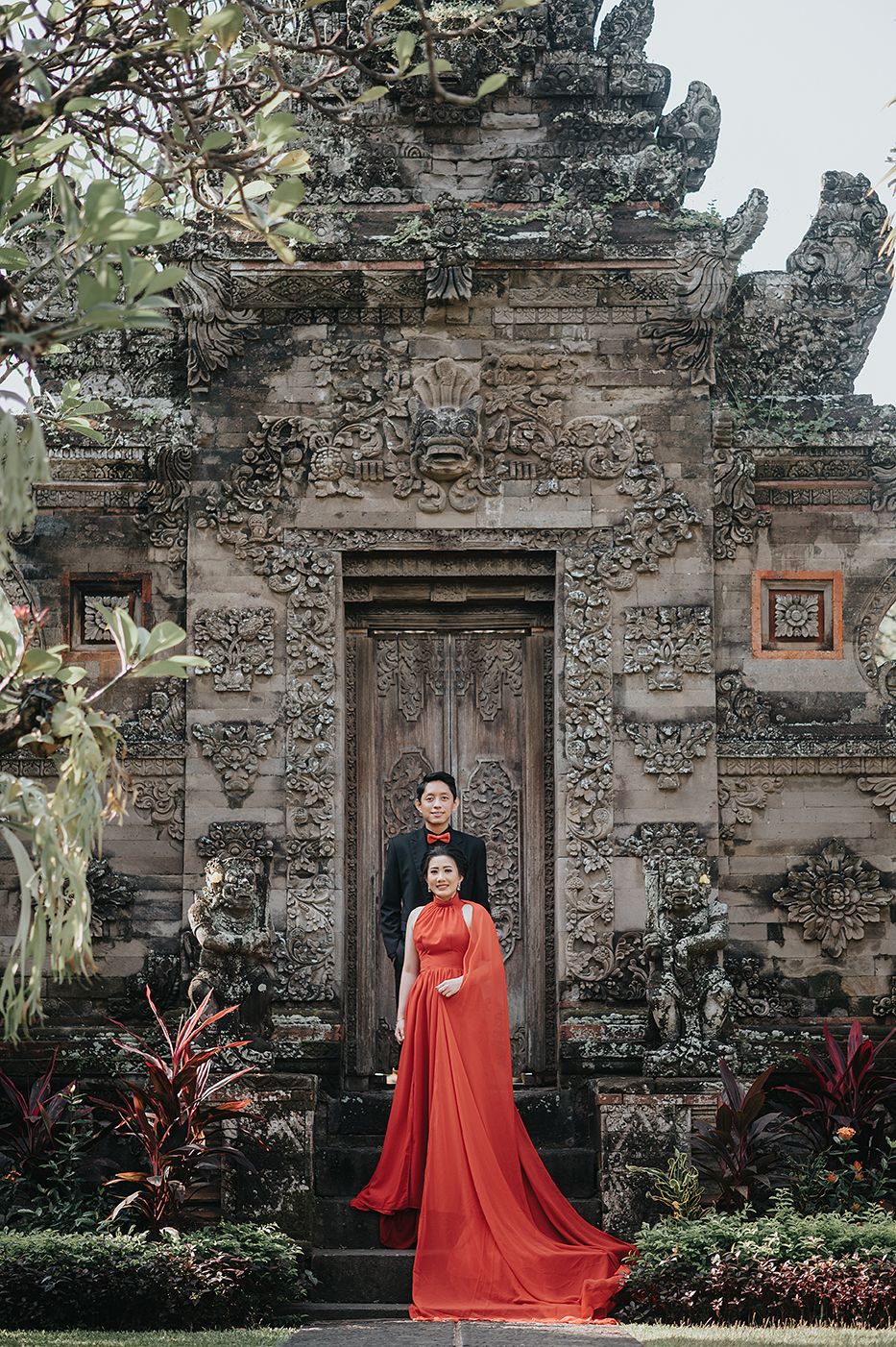 Couple posing in front of ornate Balinese temple gate, woman in flowing red gown, man in black suit.