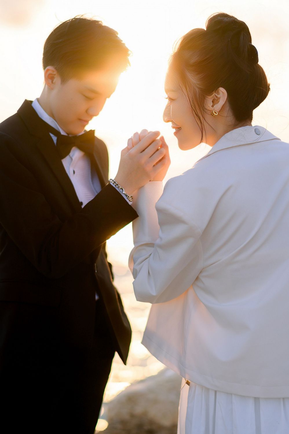 A couple in formal attire shares a romantic moment at golden hour, the groom kissing the bride's hand tenderly.