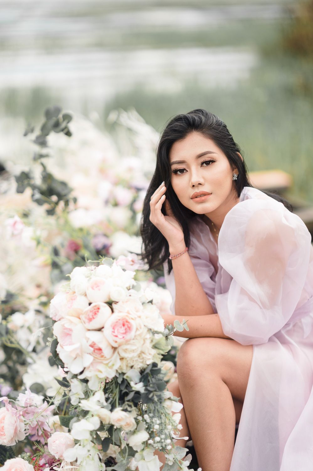 Young Asian woman in sheer pink robe poses gracefully among lush white and blush floral arrangements outdoors.