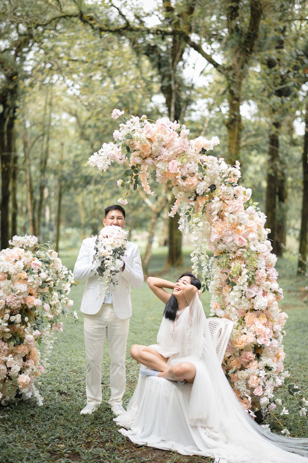 Couple in white attire poses under a floral arch with pink blooms in a lush forest wedding setting.