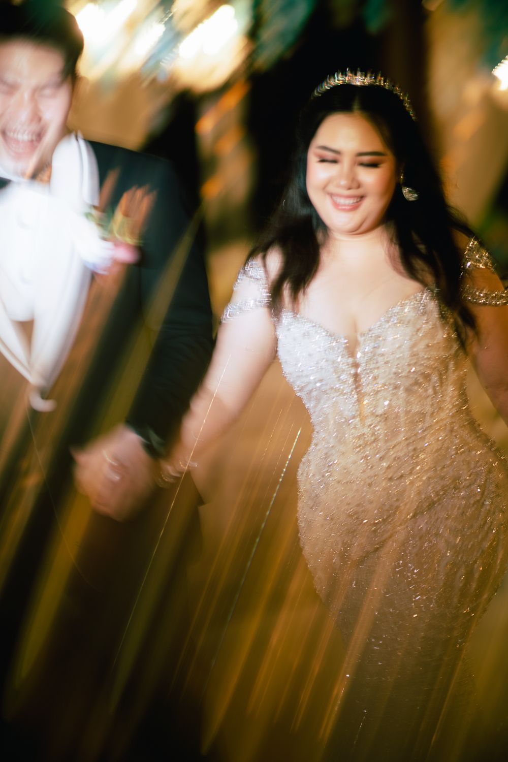 Bride in beaded gown and crown smiles joyfully with groom in tuxedo during wedding reception with golden bokeh lights.