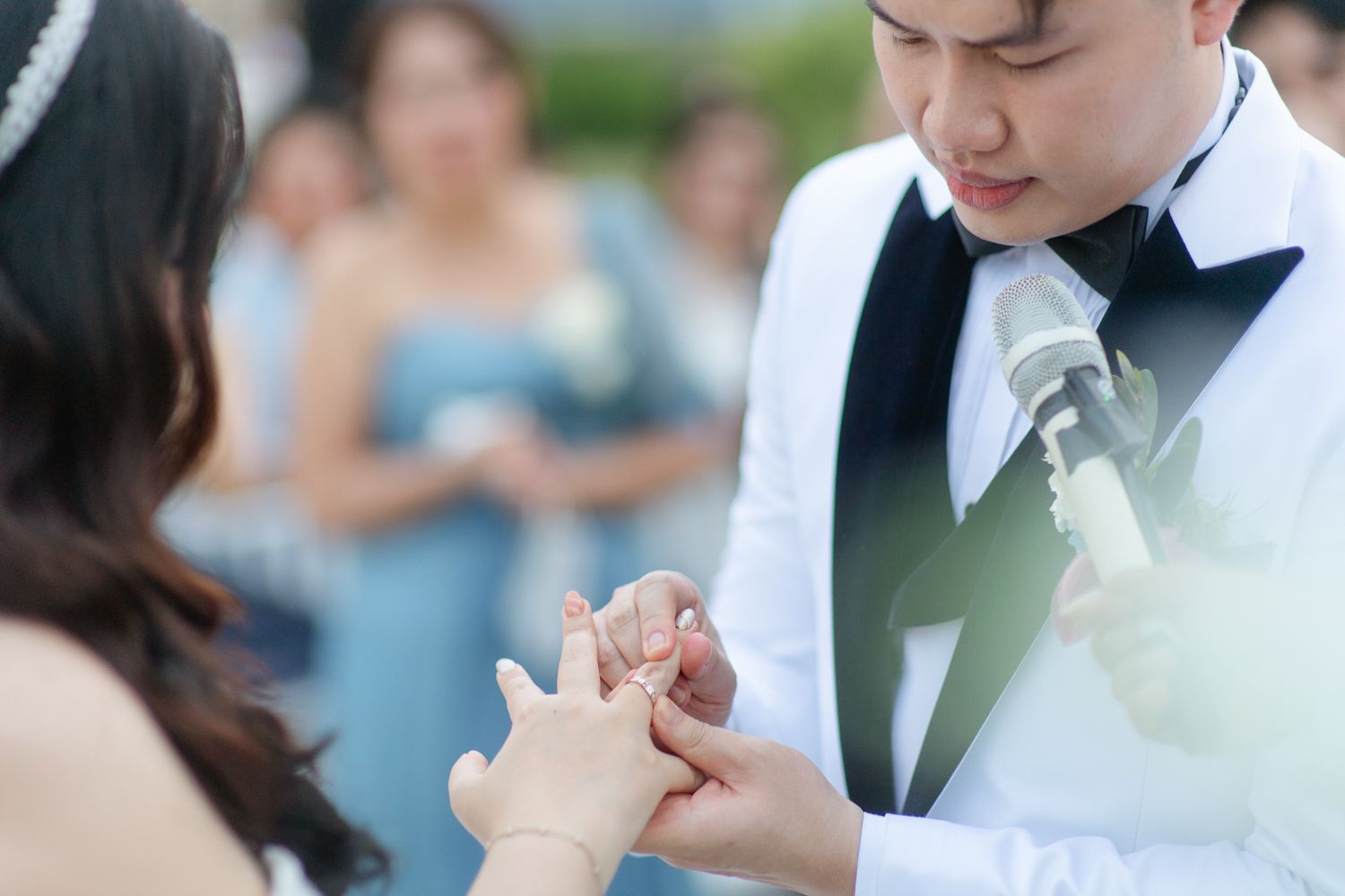 Groom in white tuxedo placing wedding ring on bride's finger during outdoor ceremony with guests in background.