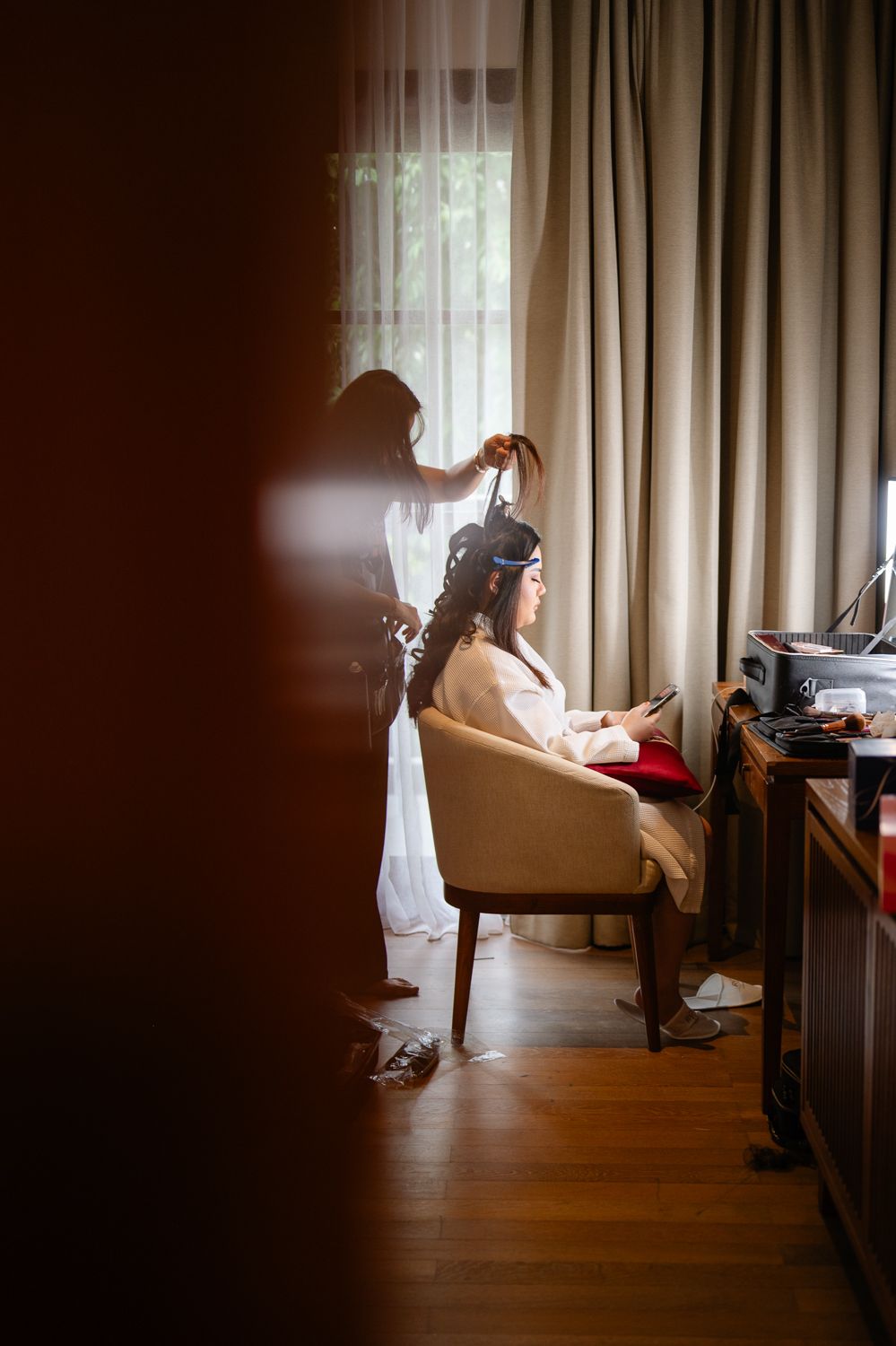 Bride sitting in chair getting hair styled by hairdresser near window with curtains in warmly lit room.