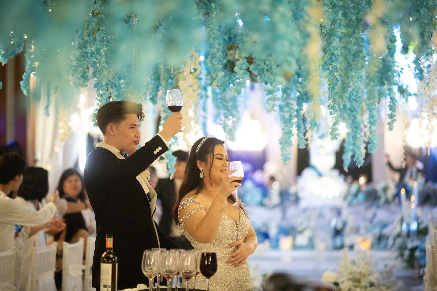 Bride and groom toasting with wine glasses at elegant blue-themed wedding reception with floral ceiling decorations.