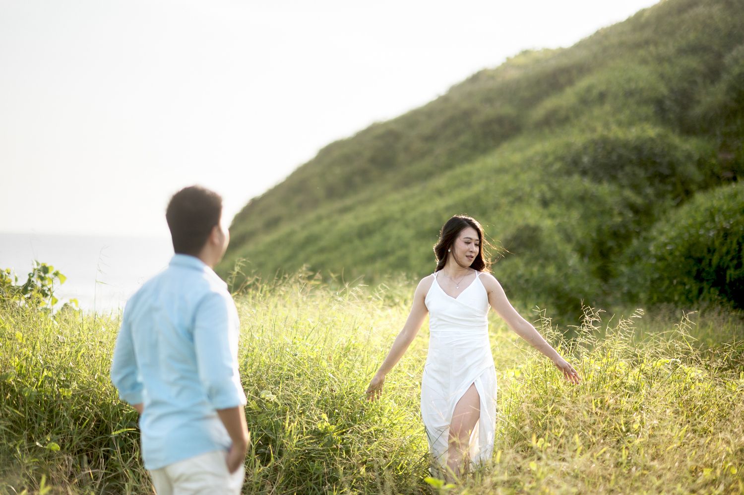 Couple walking through lush green meadow, woman in white dress smiling back at man in light blue shirt.