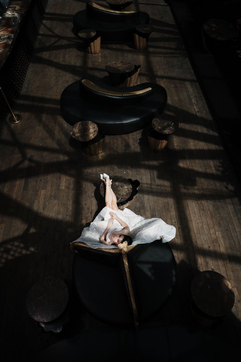 Aerial view of a bride in white dress lying on a chair in a dark interior with dramatic shadows and wooden furniture.
