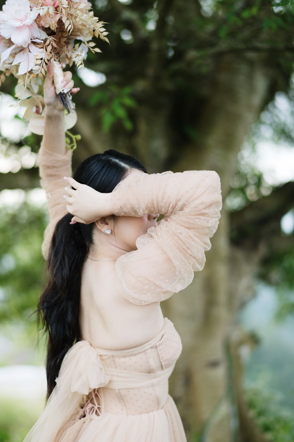 Woman in blush tulle dress reaching up to touch floral arrangement hanging from tree outdoors.