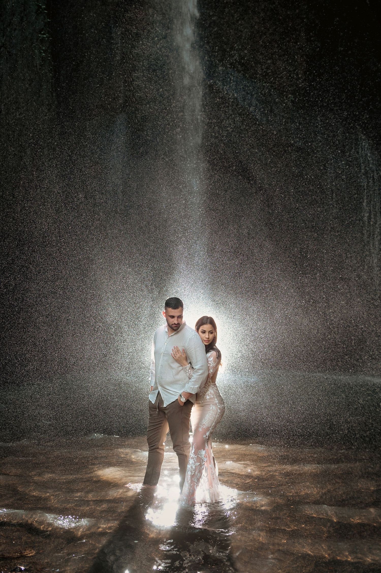 Couple standing together under dramatic waterfall spray in glowing backlit romantic setting.