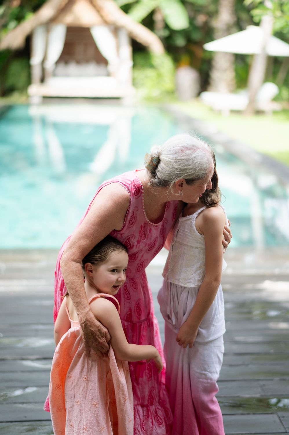 Grandmother in pink dress hugging two young girls near a tropical pool, sharing a warm family moment outdoors.