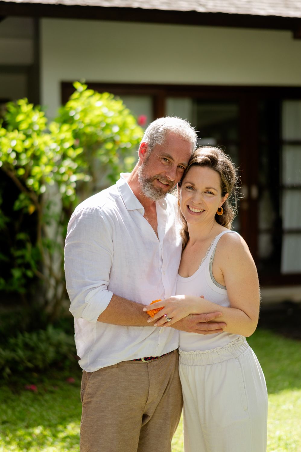 Happy middle-aged couple embracing outdoors in a sunny garden, smiling warmly in front of a tropical home.