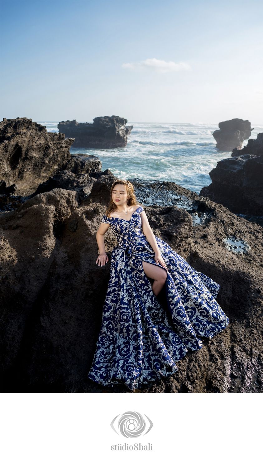 Woman in blue floral gown poses on rocky coastal cliffs with ocean waves in background, Bali photography.