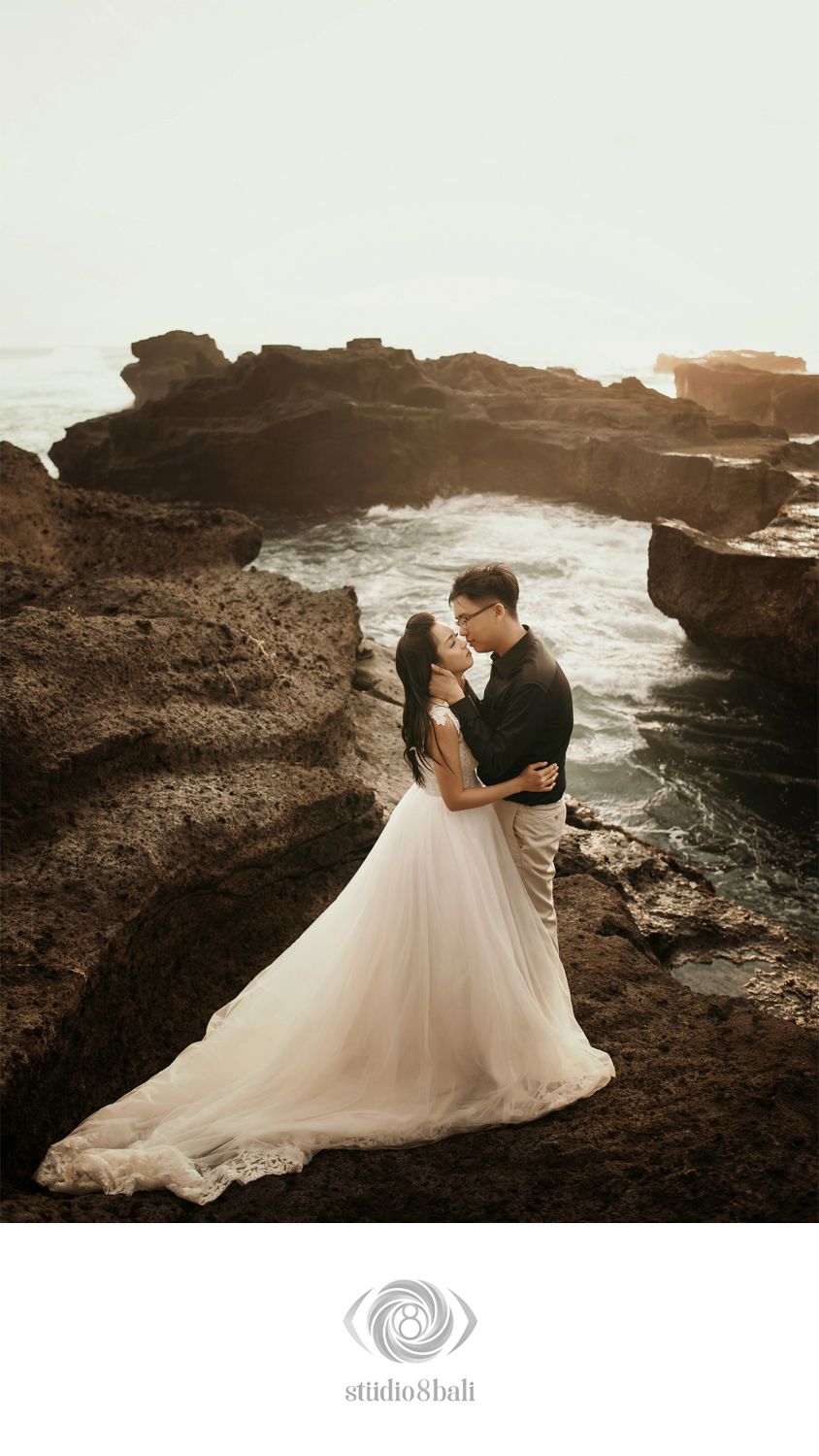 Couple in wedding attire embracing on rocky coastal cliffs with ocean waves in Bali.