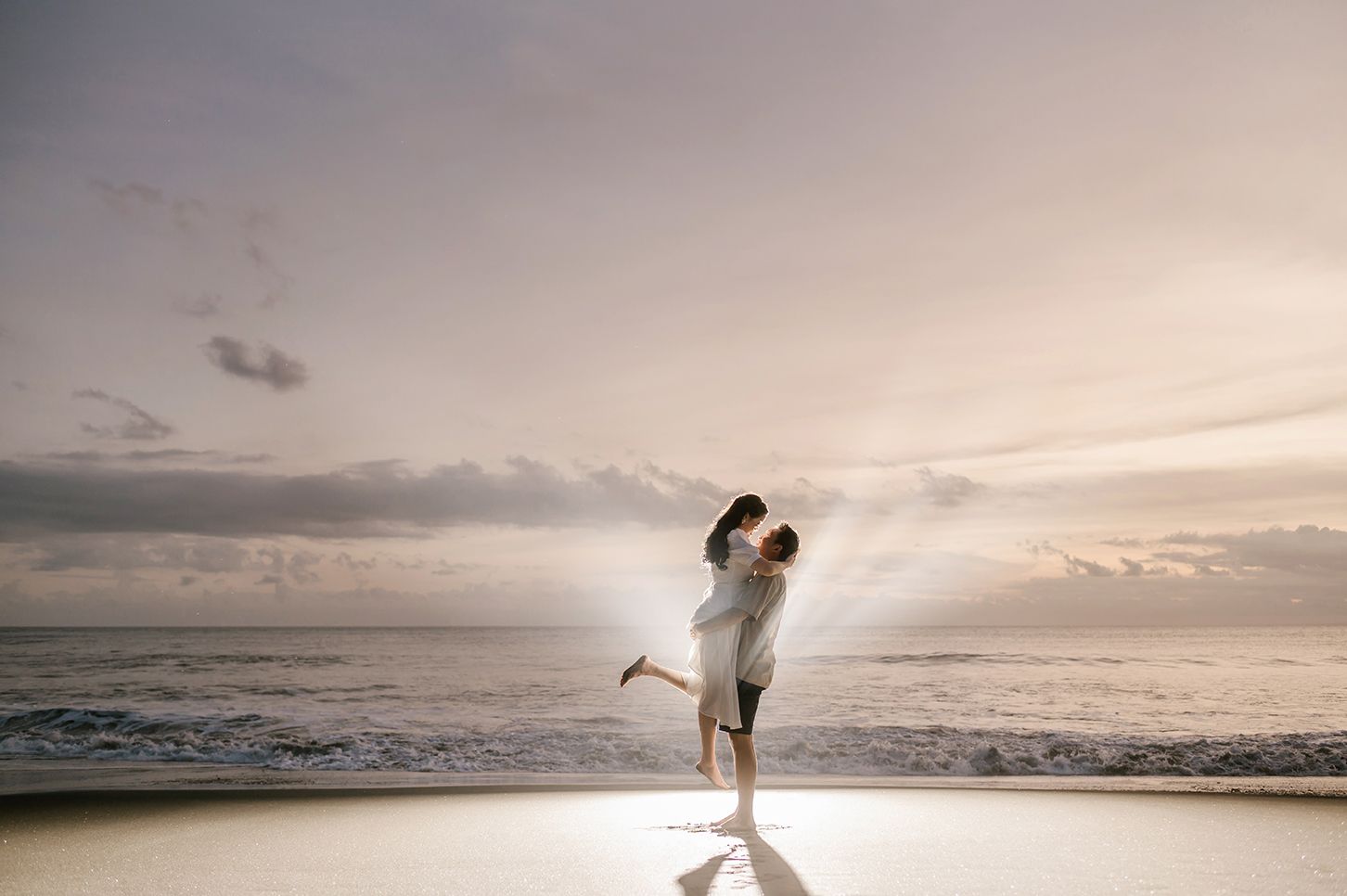 Couple embracing on a sandy beach at sunset, waves crashing behind them in a romantic seaside moment.