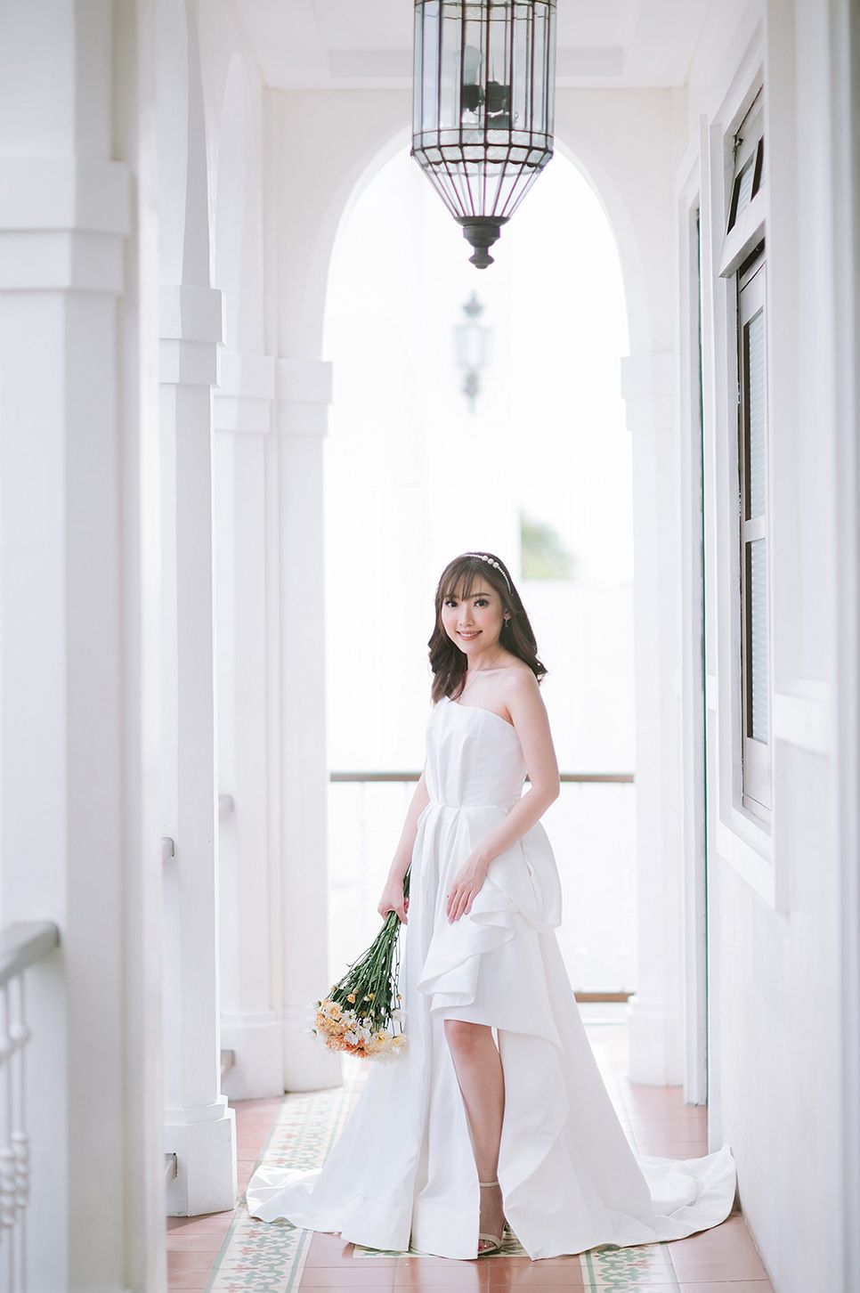 Bride in white strapless high-low wedding gown holding yellow flowers in elegant arched corridor.