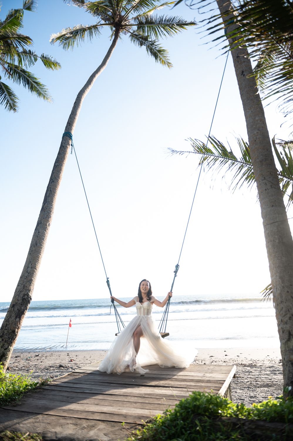 Bride in white gown sitting on a rope swing between palm trees on a tropical beach.