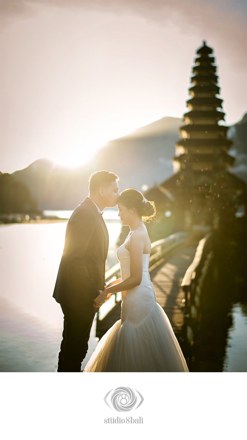 Couple sharing a romantic kiss at golden hour near a Balinese temple, bride in white gown, groom in dark suit.