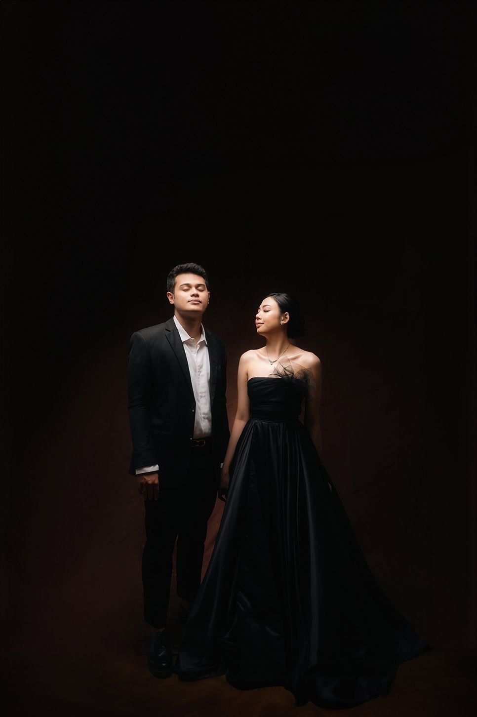 Elegant couple in black formal wear posing dramatically against dark background in a studio portrait.