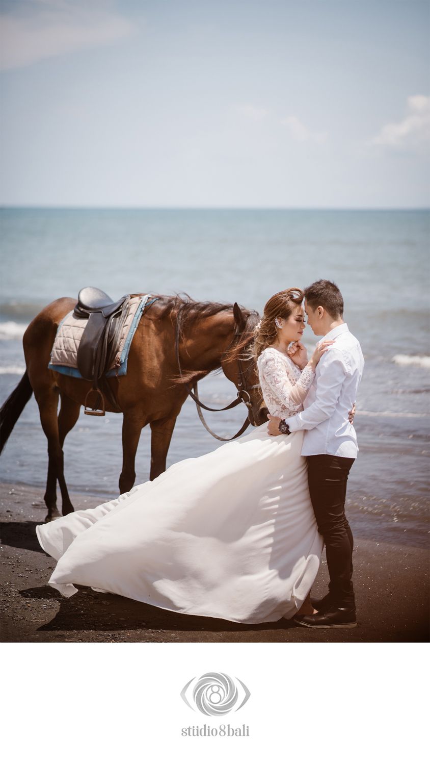 Couple in wedding attire embracing on a black sand beach beside a saddled brown horse with ocean waves behind them.