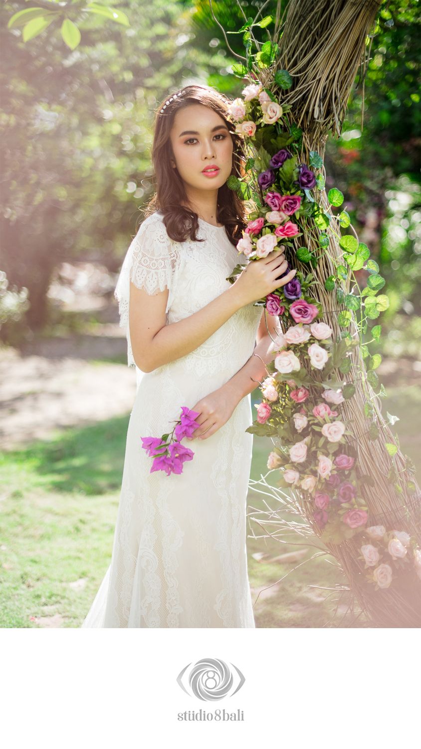 A bride in a white lace dress holds pink flowers beside a floral-adorned palm tree in a sunlit garden.