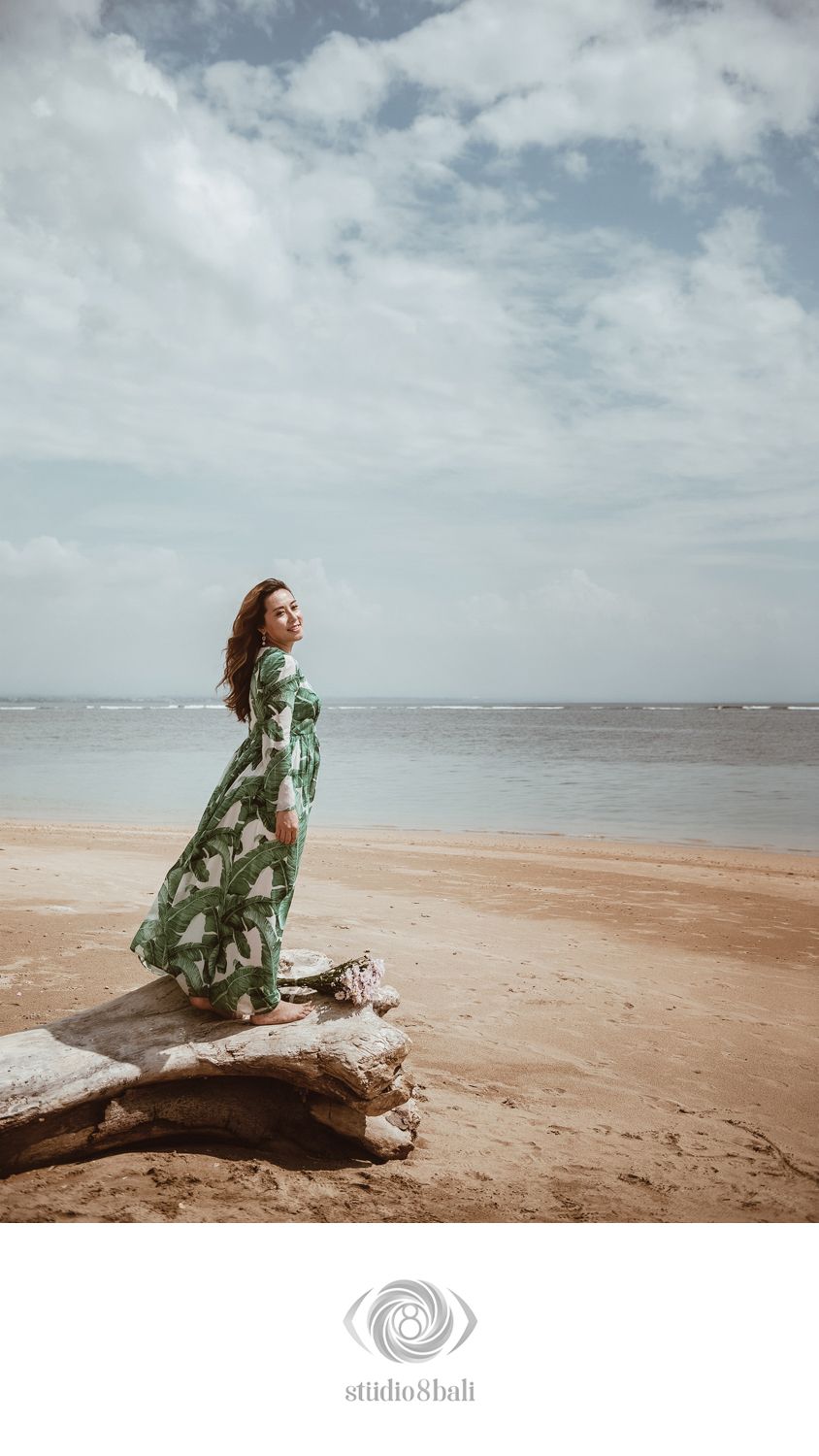 Woman in green leaf print maxi dress stands on driftwood gazing at ocean on sandy beach.