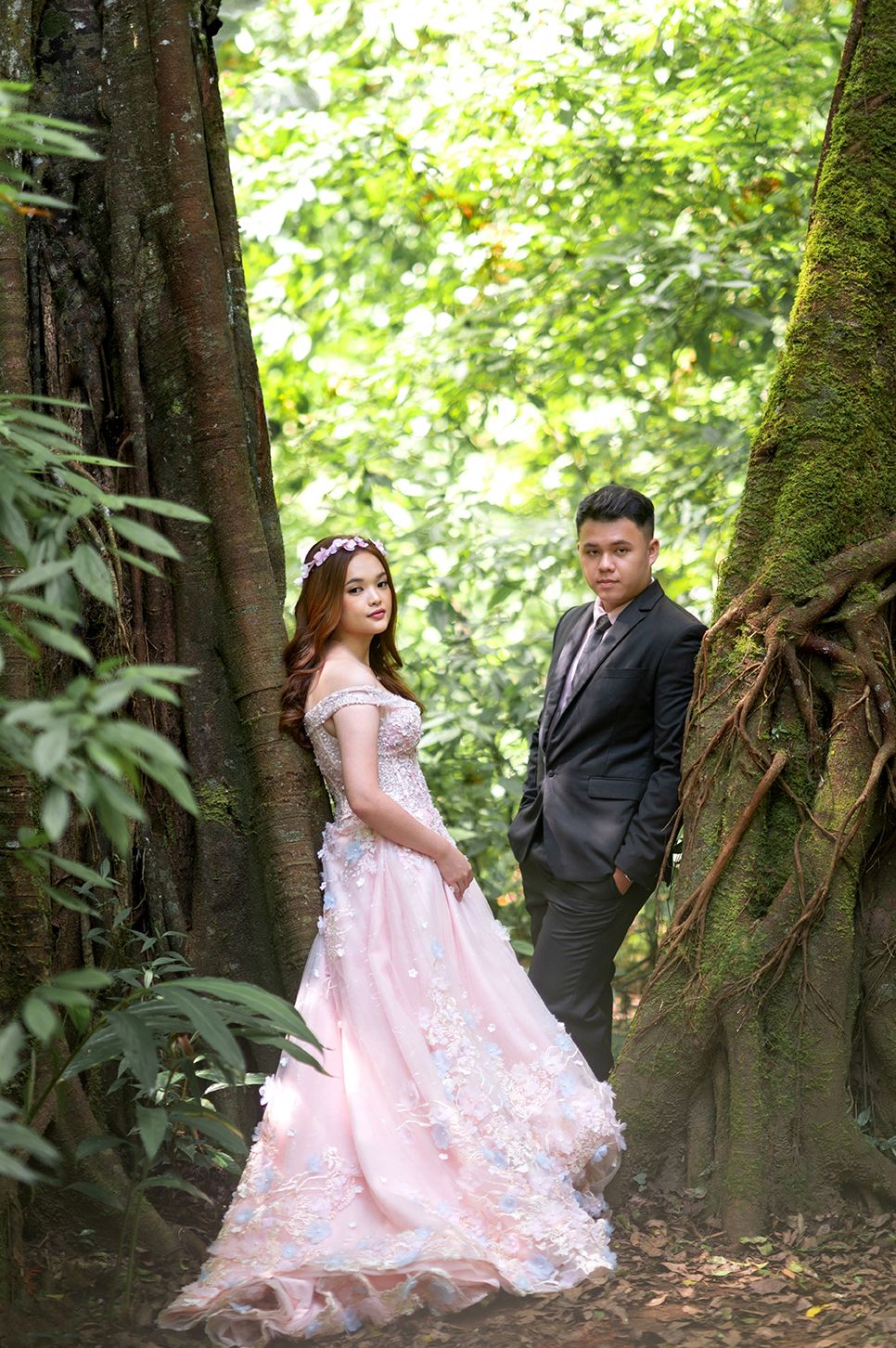 A couple poses in a lush forest, woman in pink floral gown and man in dark suit, for a romantic pre-wedding shoot.