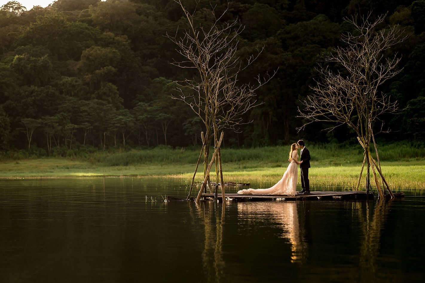 Bride and groom standing on a lakeside dock surrounded by bare trees with lush green forest backdrop at sunset.
