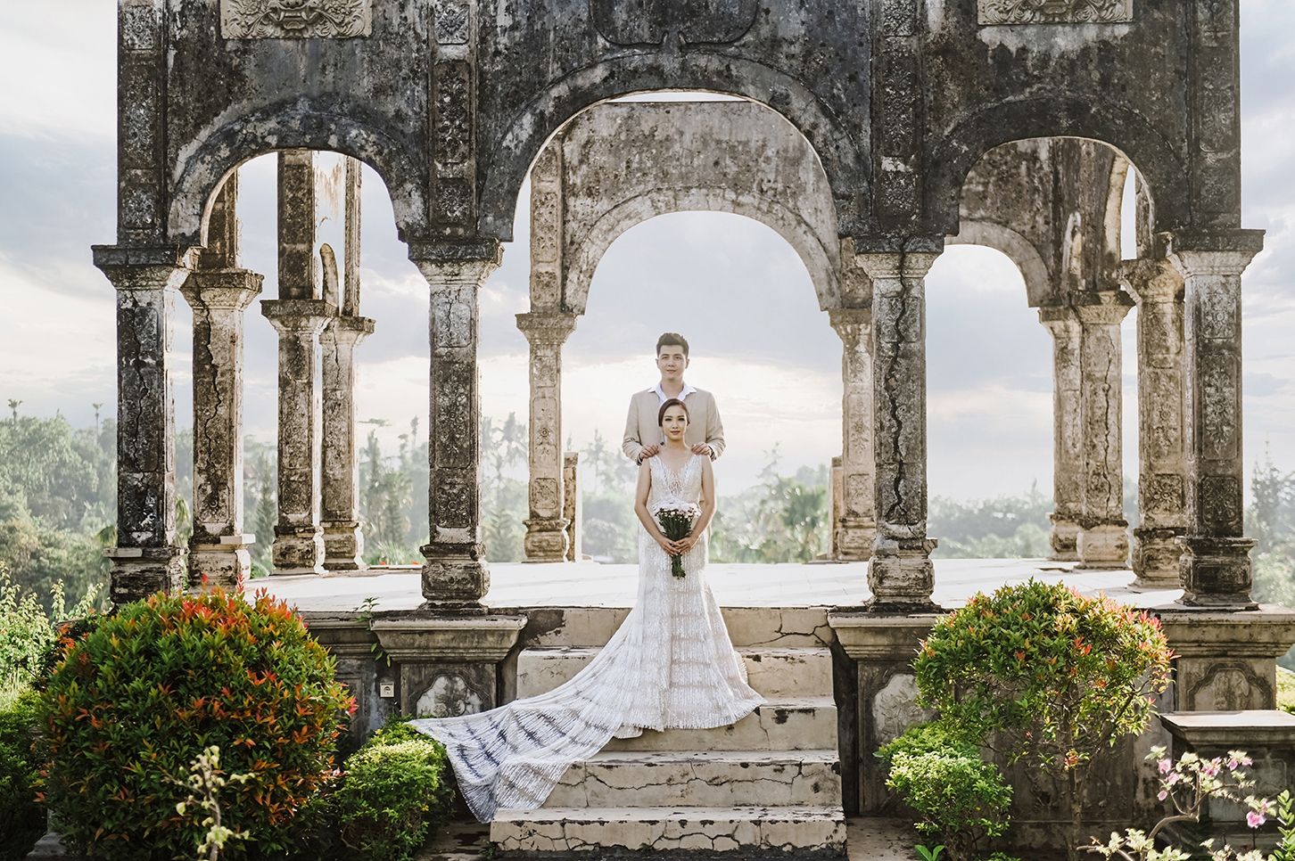 Bride and groom pose under ornate stone arches at a romantic outdoor wedding venue surrounded by lush greenery.