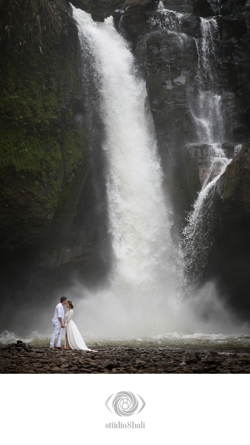Couple in white embracing before a majestic waterfall in Bali, captured by Studio 8 Bali photographers.