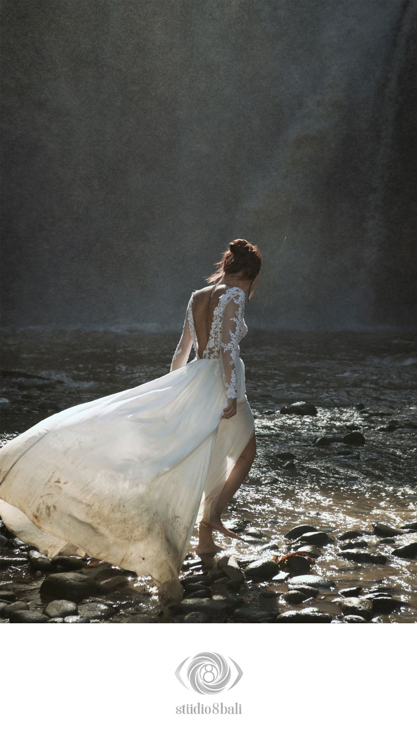 Bride in lace wedding gown standing on rocky riverbank near waterfall in Bali, dress flowing in the breeze.