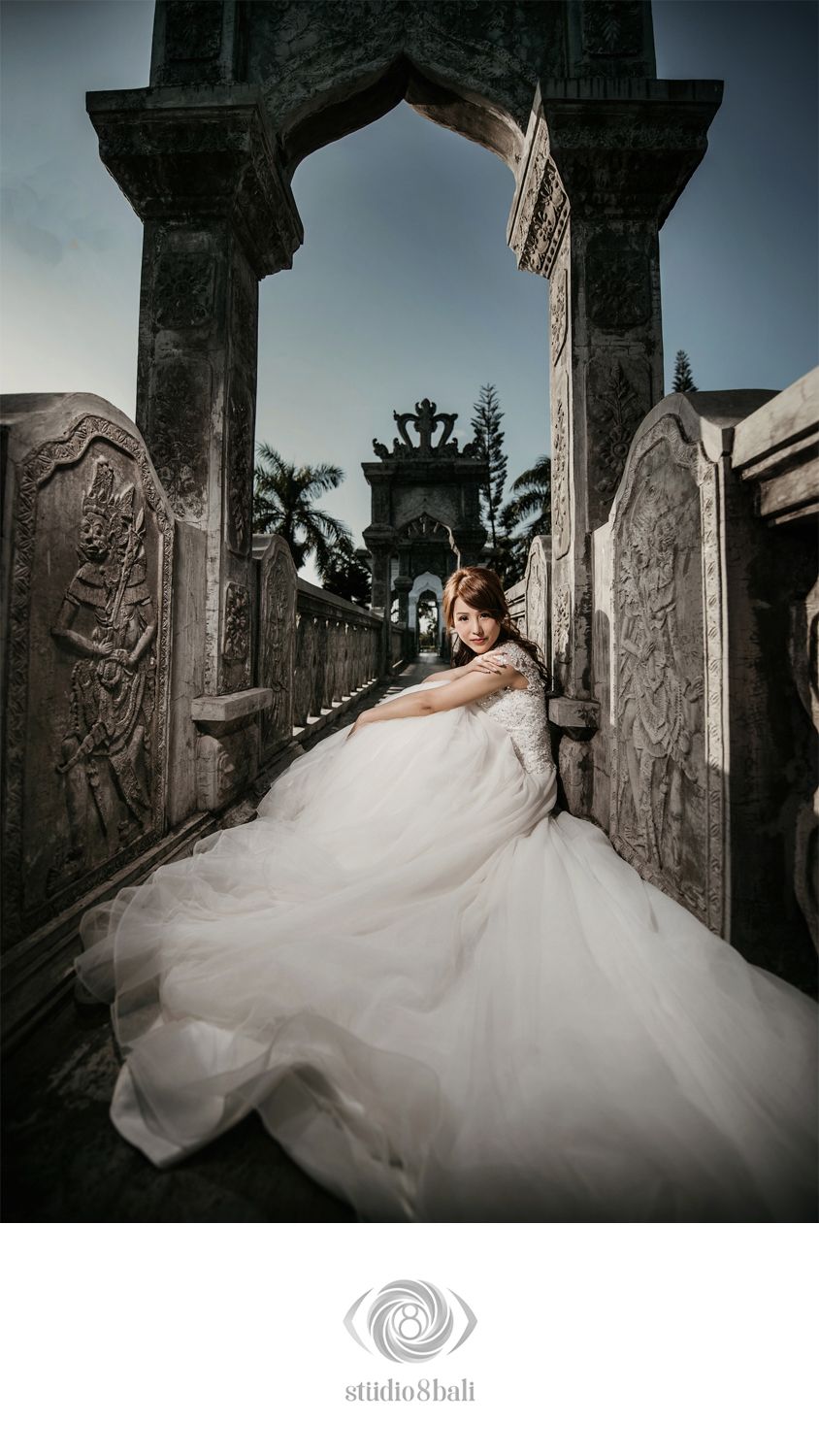 Bride in white ball gown posing at ornate Balinese stone gate temple entrance for pre-wedding photo shoot.