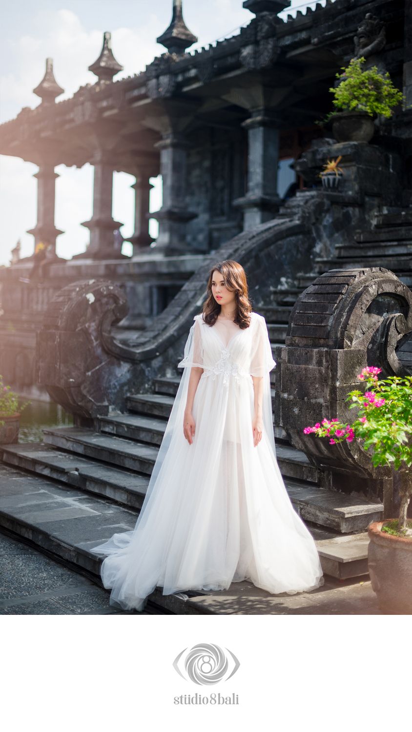 A bride in a flowing white tulle wedding gown with flutter sleeves poses before ancient stone temple steps in Bali.