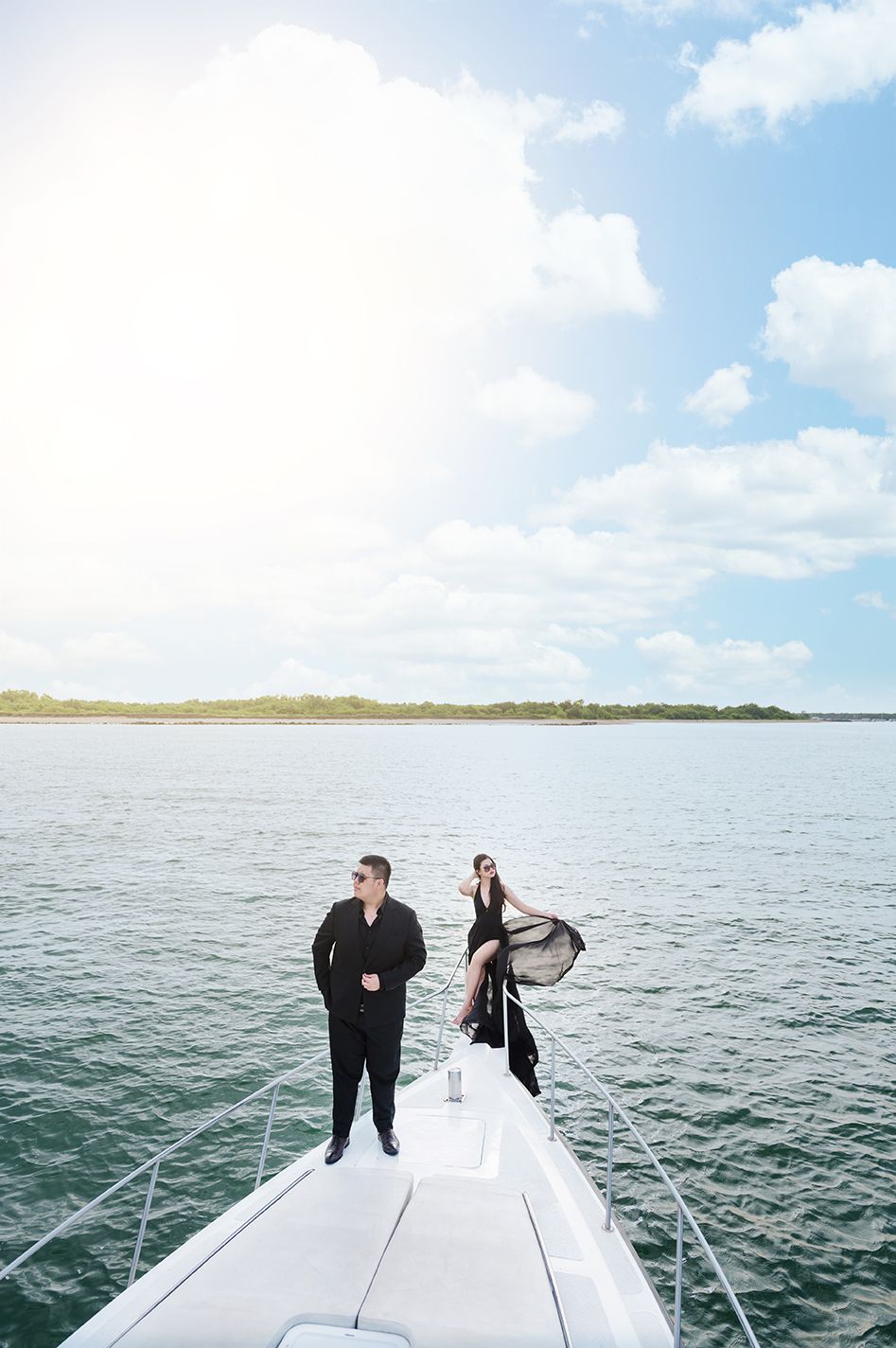 Elegant couple in black outfits posing on the bow of a luxury yacht on calm waters.