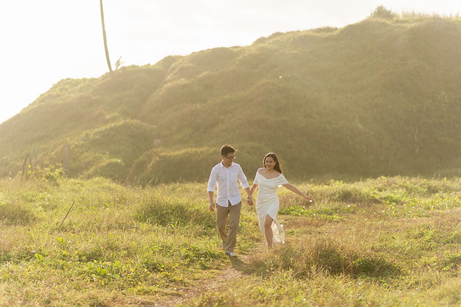 A couple holds hands and walks through a sunlit grassy field with a green hill in the background.